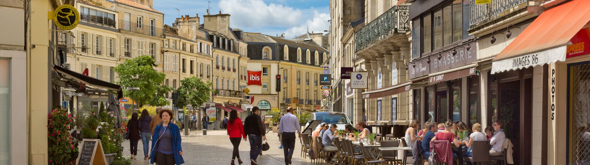 Lively street in a French city with Haussmann-style buildings, shops, a bustling café terrace with customers, and pedestrians walking on a cobbled square on a sunny day.