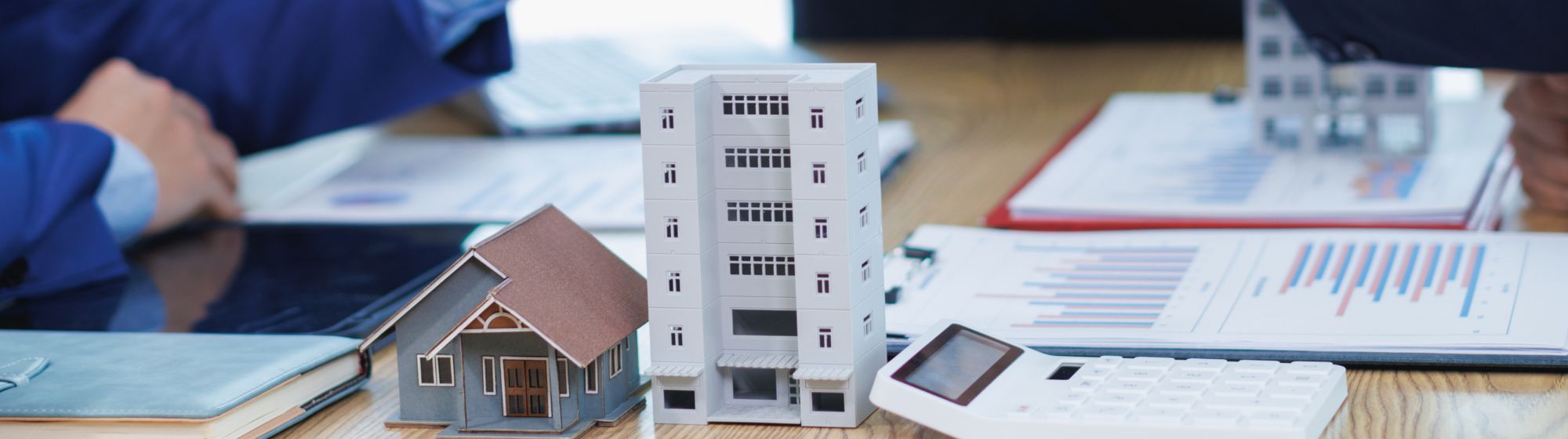 Maquettes d’une maison et d’un immeuble posées sur une table avec des documents financiers, une calculatrice et des graphiques, pendant une réunion d’investissement immobilier.