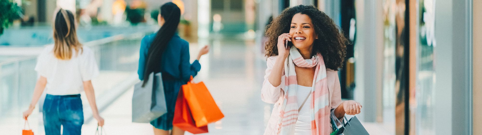 Smiling woman walking through a bright shopping mall, carrying multiple shopping bags and talking on the phone. In the background, other shoppers are also carrying bags of various colors. The image represents a dynamic and connected shopping experience.