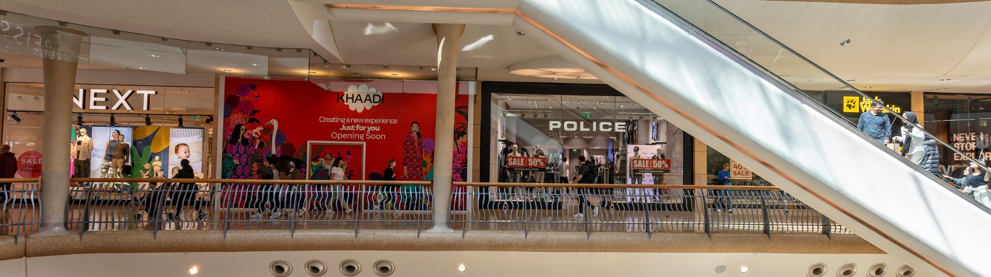 Vue intérieure d'un centre commercial avec plusieurs boutiques, dont Next, Police et Jack Wolfskin, et un panneau d’ouverture prochaine pour la marque Khaadi. Des clients marchent et font leurs achats sous un escalier moderne.