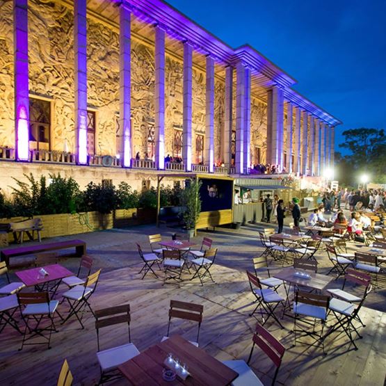 Soirée de gala en extérieur au Palais de la Porte Dorée à Paris, avec des tables et chaises en bois disposées sur une terrasse, une façade illuminée de lumières violettes et dorées, et des invités profitant de l'événement sous un ciel nocturne.