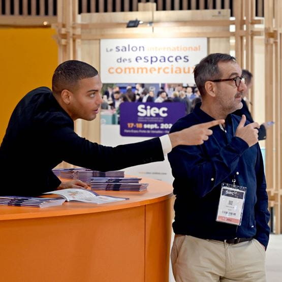 Un membre de l’équipe d’accueil du salon Siec aide un visiteur en lui indiquant une direction, devant un comptoir d'information orange et une affiche du salon en arrière-plan.
