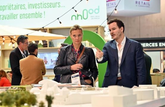 A professional presents an architectural model to a visitor at a trade show, showcasing urban development projects.