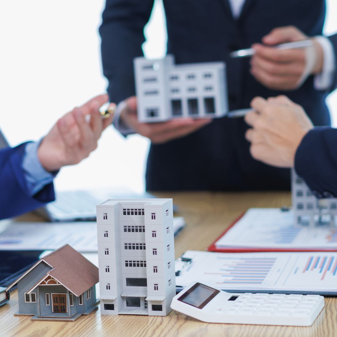 Real estate investment meeting with building models, a calculator, and financial documents on a table, while men in suits discuss in the background.