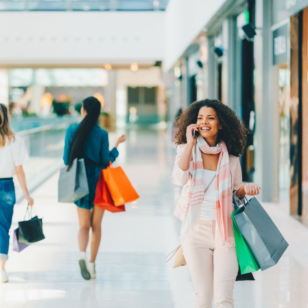 Smiling woman walking through a bright shopping mall, carrying multiple shopping bags and talking on the phone. In the background, other shoppers are also carrying bags of various colors. The image represents a dynamic and connected shopping experience.