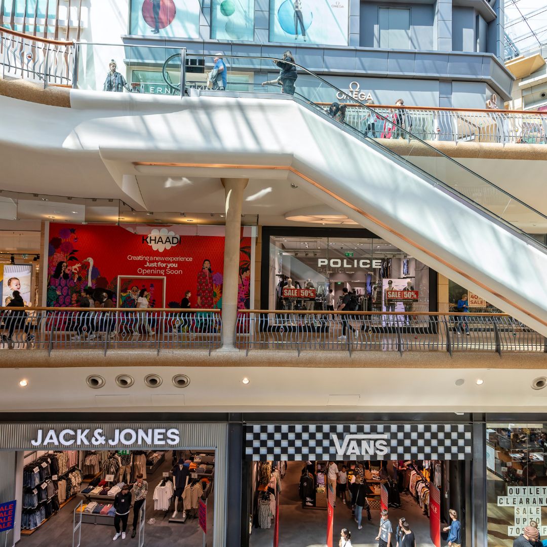 Interior view of a modern shopping mall featuring stores like Jack & Jones, Vans, Police, and Khaadi. Shoppers walk and browse under a stylish staircase.